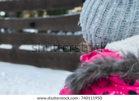 girl in gray cap and pink jacket standing back against wooden background brown fence and pure snow