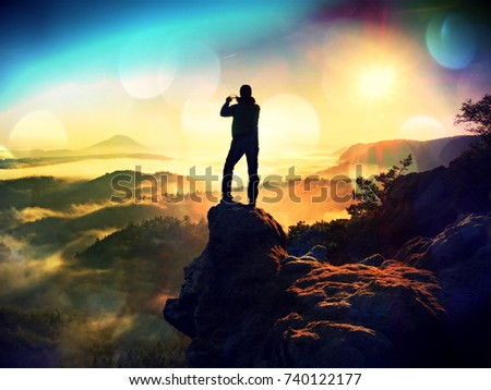 Flare, soft focus.Tall backpacker with phone in hand makes selfie. Sunny spring daybreak in rocky mountains. Hiker with backpack stand on rocky view point