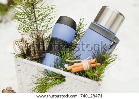 Thermocup, thermos with tea and pine branches lying in a basket on a white snowy background in winter season. Winter concept. Blue thermos