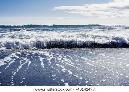 Little waves of Pacific ocean are broken on the shore. Splashes of it goes on a black volcano sand surface of a beach. Kamchatka. Russian Federation. October of 2017. Light blue sky.