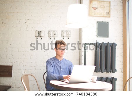 Young man writing a business plan for coffee shop on laptop computer. Handsome hipster guy keyboarding on portable net-book, sitting in cafe. Handsome male manager typing text on modern notebook