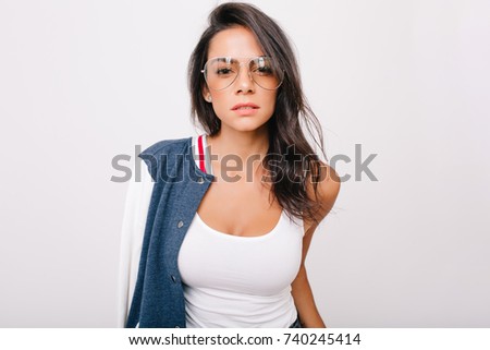 Close-up portrait of stunning dark-haired girl in white tank-top looking with interest to camera. Indoor photo of adorable european lady in stylish glasses chilling on photoshoot.