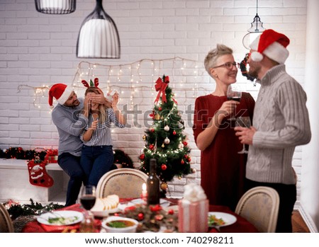 Young happy girl sitting on the boyfriend's leg while he holding her eyes closed for Christmas party.