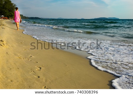 a young girl in a pink dress walks barefoot along the beach of Cambodia, along the seashore, leaving traces on the sand. the wave foams
