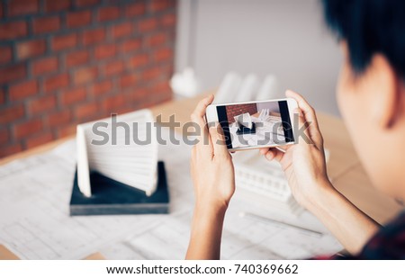 Architect man holding smartphone taking a photo of an architecture model sent to client and working in home office.