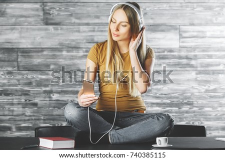 Portrait of modern young european woman taking picture with smartphone and listening to music while sitting on top of office desk with coffee cup and other items. Music concept 