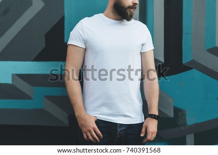 Summer day. Front view. Young bearded hipster man dressed in white t-shirt is stands against wall with graffiti. Mock up. Space for logo, text, image.