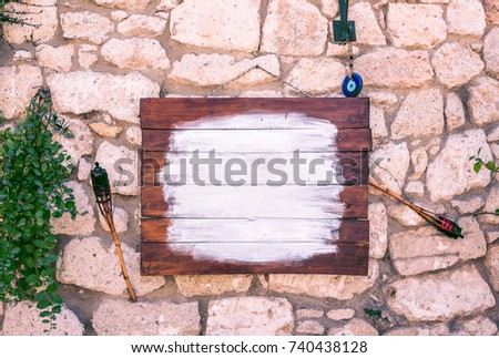 Front view of free and clean brown wooden signage board on a white ancient stone wall with hanging plants and evil eye bead for decoration.There is a copy space for editing.
