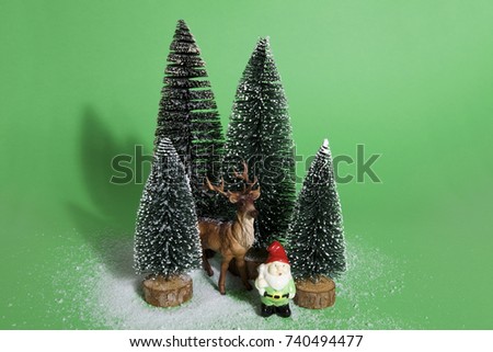 Isolated group of full artificial firs like a small forest tree in a ceramic pattern plate with a figurine reindeer and garden gnome beside on a green background. Minimal still life photography