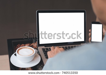 Casual business man working on white blank screen laptop computer, browsing internet and using mobile smart phone with cup of coffee on table at coffee shop, working at cafe, mock up image, close up