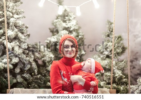 Happy family mother and child boy in santa hat near snow covered Christmas trees with sitting on swing. Bright garland above heads