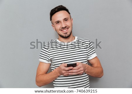 Picture of smiling young man chatting by mobile phone isolated over grey background. Looking camera.