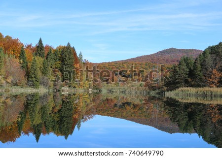 A mountainous forest with autumn colors is reflected in a clear pond. The lake is serene under a blue sky. The fall scene is from Croatia's beautiful Plitvice Lakes National Park (Plitvi?ka Jezera).