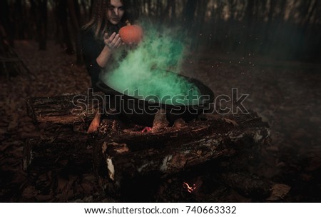 A beautiful girl witch cooking an infernal potion in a cauldron at the stake in a dark forest. The Art of Halloween