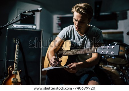 Handsome young man on rehearsal base. Lyric singer with acoustic guitar is writing notes and lyrics in notebook. Royalty-Free Stock Photo #740699920
