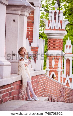 A blonde girl in a beige dress and lace shawl standing by a red brick wall in a park with a bouquet of flowers and smiling. Soft sunlight. Summer time 