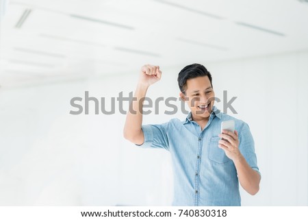 Happy excited Asian man looking at his smartphone and raising his arm up to celebrate success or achievement. Royalty-Free Stock Photo #740830318