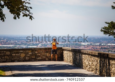 the tourist man stands with his back on the viewing platform in Bergamo in Italy and takes a photo on the camera wearing shorts, sandals and an orange backpack. General view of the city