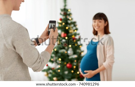winter holidays, technology and people concept - happy husband photographing his pregnant wife by smartphone at home at christmas
