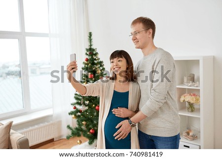 winter holidays, technology and people concept - happy husband and his pregnant wife taking selfie by smartphone home at christmas