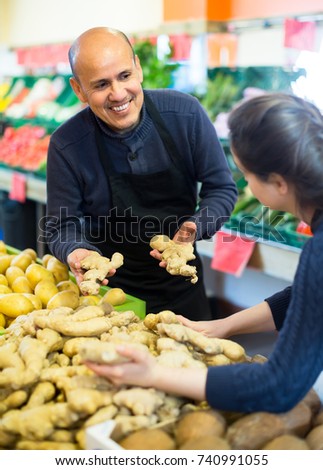 Friendly mature seller offering female fresh roots of ginger in grocery
