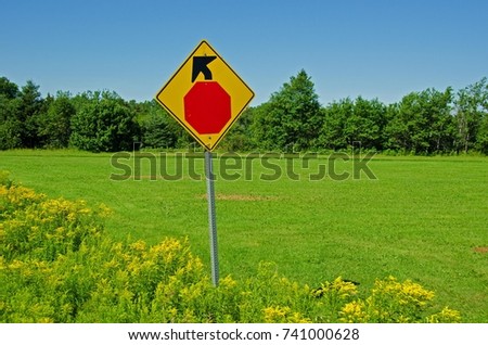 Yellow and red stop sign shown against a green farm field with blue skies and line of trees in background.