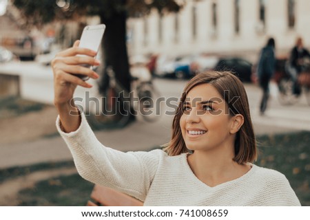 Young beautiful brunette taking pictures of herself on a cellphone in a city park.