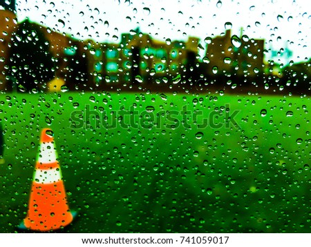 Image of raindrops on the clear window with blurred school soccer field and blurred building in the background in a rainy day. Blurry orange soccer training cone is on the left corner.