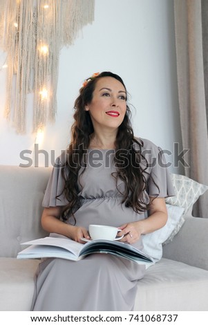 pregnant woman sits in a chair with a cup of tea and with a glossy magazine lights on the background