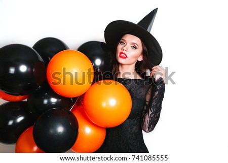 Portrait of beautiful young woman in black witch halloween costume with orange and black balloons over white background