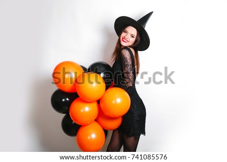 Portrait of beautiful young woman in black witch halloween costume with orange and black balloons over white background