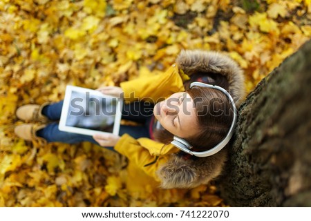 The brown-haired woman in yellow coat and jeans sitting and listening to music under a tree with a tablet in her hands and headphones in fall city park on a warm day. Autumn golden leaves. Top view