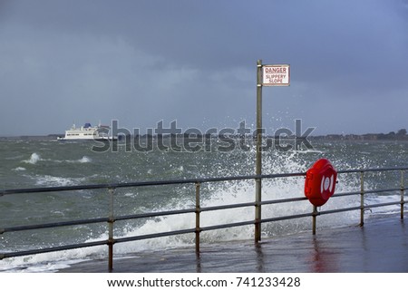 Stormy weather and heavy seas causing problems for a small ship as it passes a warning sign and life preserver on the beach at Southsea. Danger slippery slope on the promenade.
