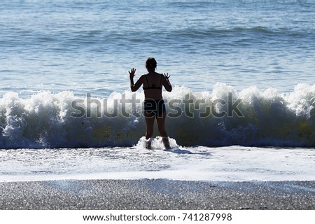 A silhouetted woman in swimwear holds up her hands as if to stop a breaking wave. Royalty-Free Stock Photo #741287998