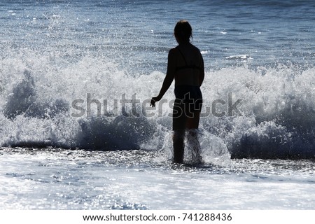 In bright sunlight the silhouette of a woman faces a breaking wave on a beach. Royalty-Free Stock Photo #741288436