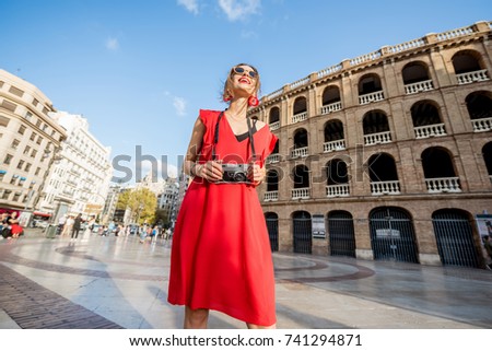 Portrait of young woman tourist in red dress standing with photo camera in front of the bullring amphitheatre in Valencia city, Spain