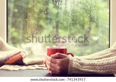 red mug of hot drink in hand, when behind is window is rain / cozy home atmosphere in autumn Royalty-Free Stock Photo #741343957