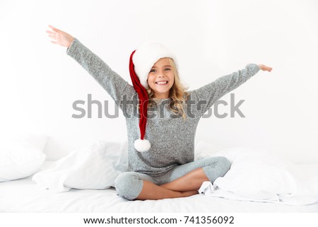 Happy exited blonde girl in Santa's hat sitting on bed with raised arms, looking at camera