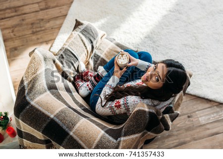top view of young woman enjoying hot chocolate with marshmallow while sitting in cozy armchair
