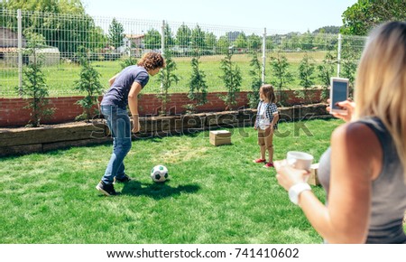 Mother taking mobile photo of her husband and son playing in the garden