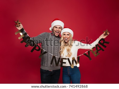 Portrait of a smiling happy couple in christmas hats holding happy new year ribbon and looking at camera isolated over red background