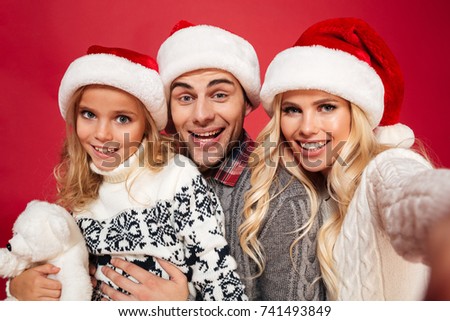 Close up portrait of a young cute family with a child wearing christmas hats while taking a selfie isolated over red background