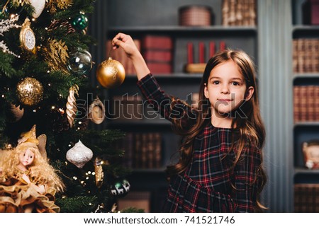 Small little kid decorates Christmas tree indoors, looks directly into camera, holds glass ball, being glad to have winter holidays. Little adorable girl wears dress decorates New Year tree alone