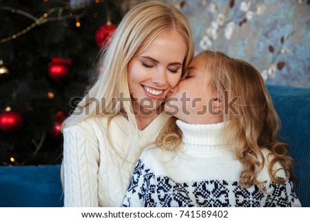 Close-up photo of little girl in knitted sweater kiss her mother while sitting on sofa at Christmas tree