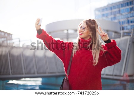 A hipster girl in a red sweater and glasses takes pictures of Selfie on the phone. Beautiful architecture in the background.
