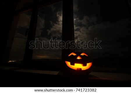 Scary Halloween pumpkin in the mystical house window at night or halloween pumpkin in night on room with blue window. Symbol of halloween in window. Selective focus