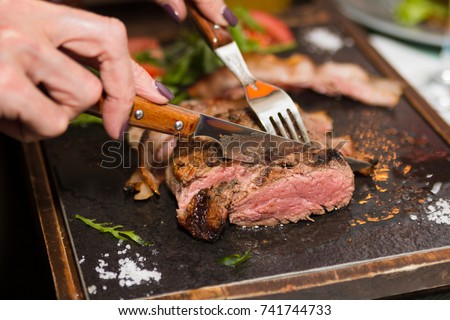 Woman hand holding knife and fork cutting grilled beef steak on stoned plate. Selective focus Royalty-Free Stock Photo #741744733