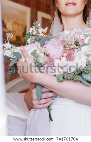 Beautiful wedding bouquet in hands of the bride. Trendy and modern wedding flowers. Peonies and roses. Woman in wedding dress in hotel room. Blurred background.