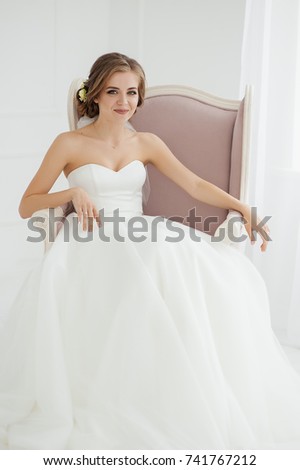 The bride is gathering in the morning. She sits in a beautiful chair. Stylish white wedding dress. 