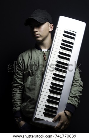 serious teen with cap and bomber jacket holding keyboard instrument in front of black background
